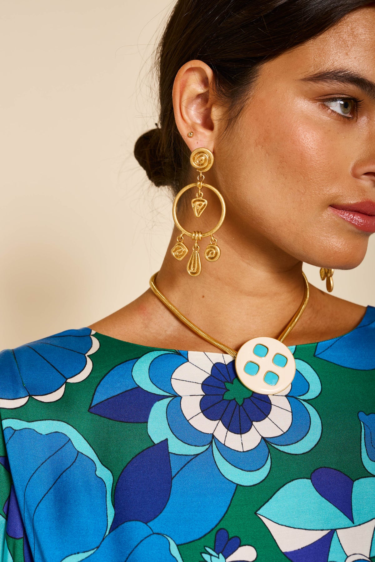 Close-up of a woman in the marni - miami floral emerald top, gold dangling earrings, and a gold necklace with a round pendant featuring four turquoise squares. Only the right side of her face and shoulder are visible.