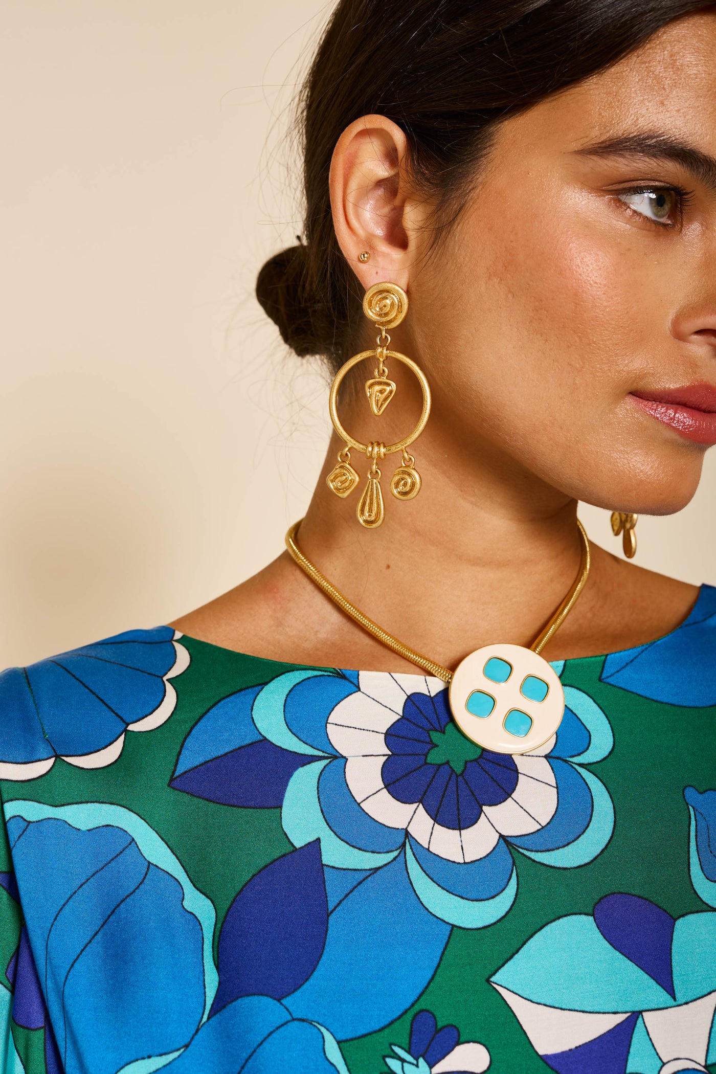 Close-up of a woman in the marni - miami floral emerald top, gold dangling earrings, and a gold necklace with a round pendant featuring four turquoise squares. Only the right side of her face and shoulder are visible.