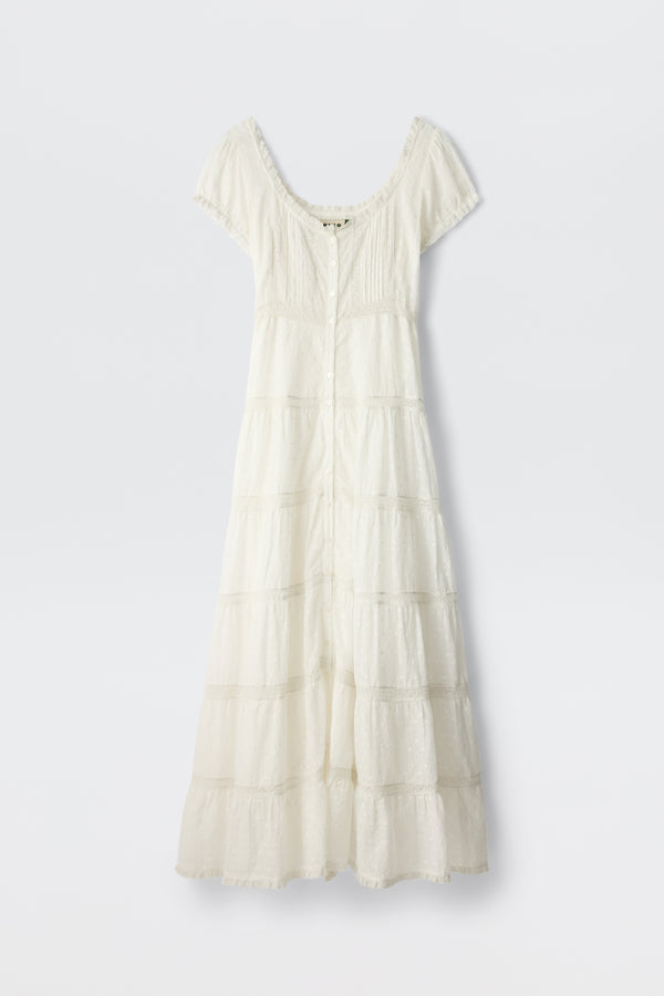 The Jaclyn - Ivory is a white cotton summer dress with short sleeves, an ankle-length tiered skirt, fitted bodice, button-down front, and lace-trimmed neckline and sleeves. The delicate fabric has a textured broderie anglaise pattern.