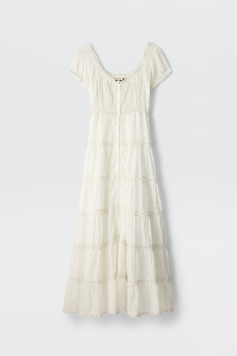 The Jaclyn - Ivory is a white cotton summer dress with short sleeves, an ankle-length tiered skirt, fitted bodice, button-down front, and lace-trimmed neckline and sleeves. The delicate fabric has a textured broderie anglaise pattern.