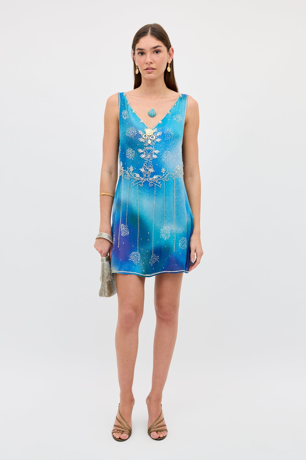 A woman wearing the Kaylor - Jessica Alexandra Blue, a sleeveless mini dress featuring blue ombré hues with silver embellishments and delicate Aari embroidery, accessorized with a small silver bag, gold sandals, and gold jewelry, stands against a plain white background.