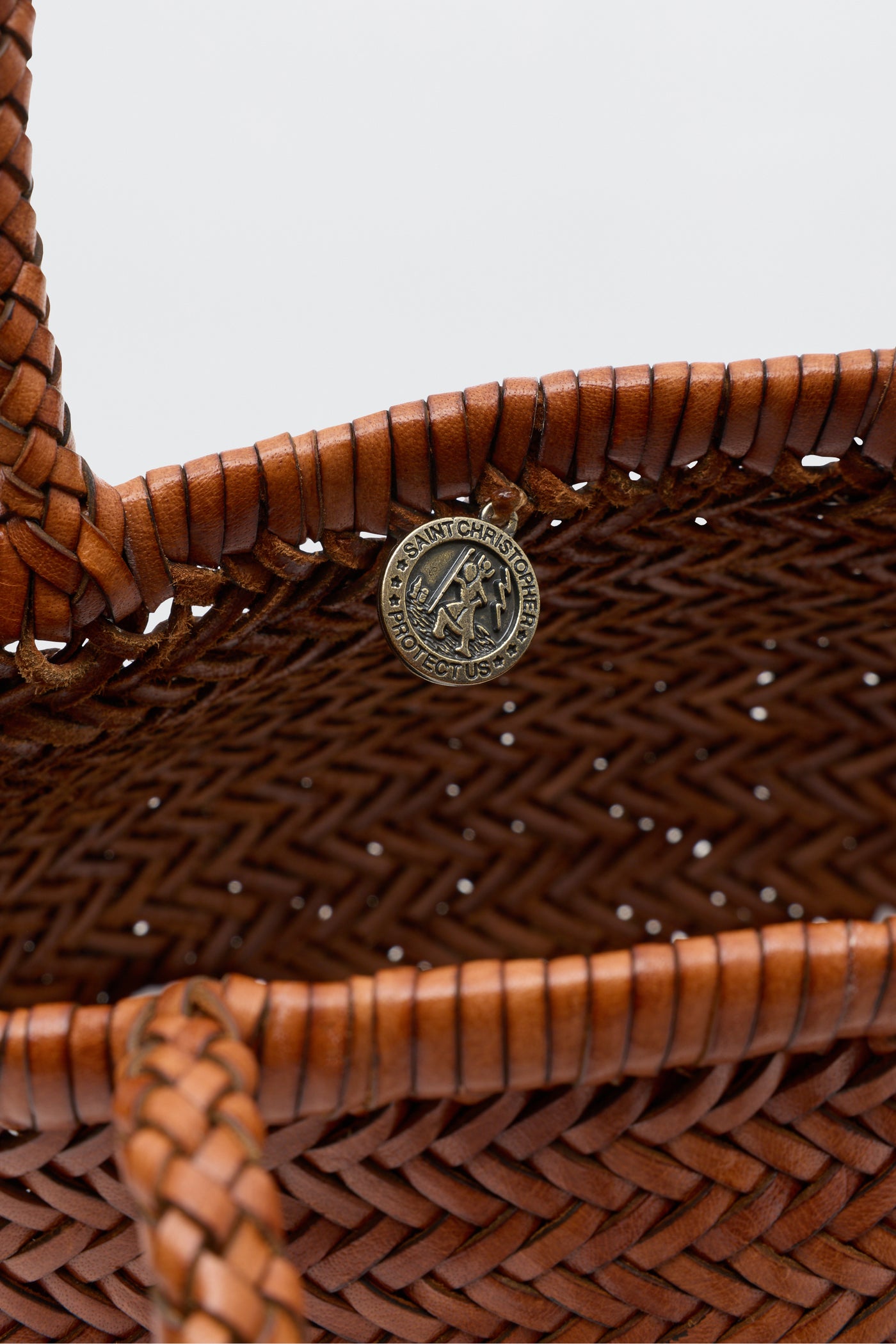 Close-up of the Lysia - Tan, a brown woven leather basket reminiscent of vintage Dragon Diffusion bags, featuring a round metal medallion reading "Saint Christopher Protect Us" attached to the inner rim.